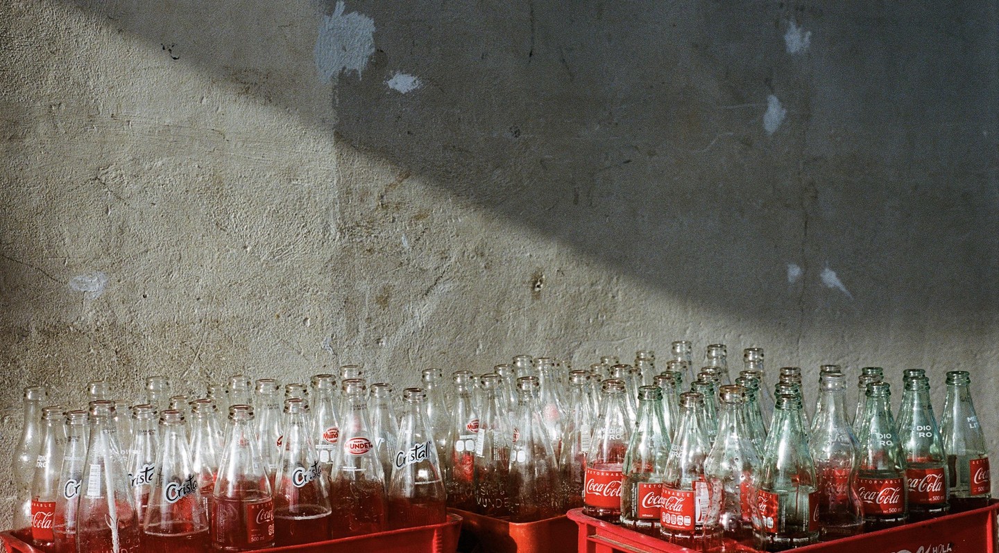 Many glass bottles of Coca-Cola and Cristal soda are arranged in red crates against a textured wall.