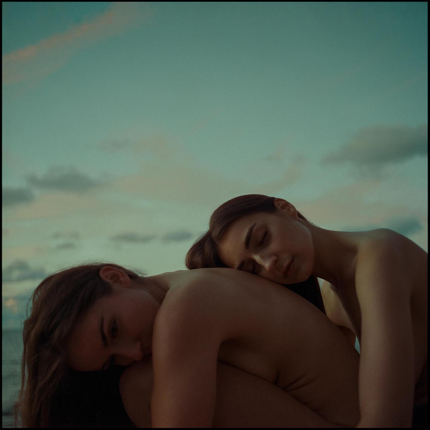 Two women are embracing each other with their eyes closed under a cloudy sky.