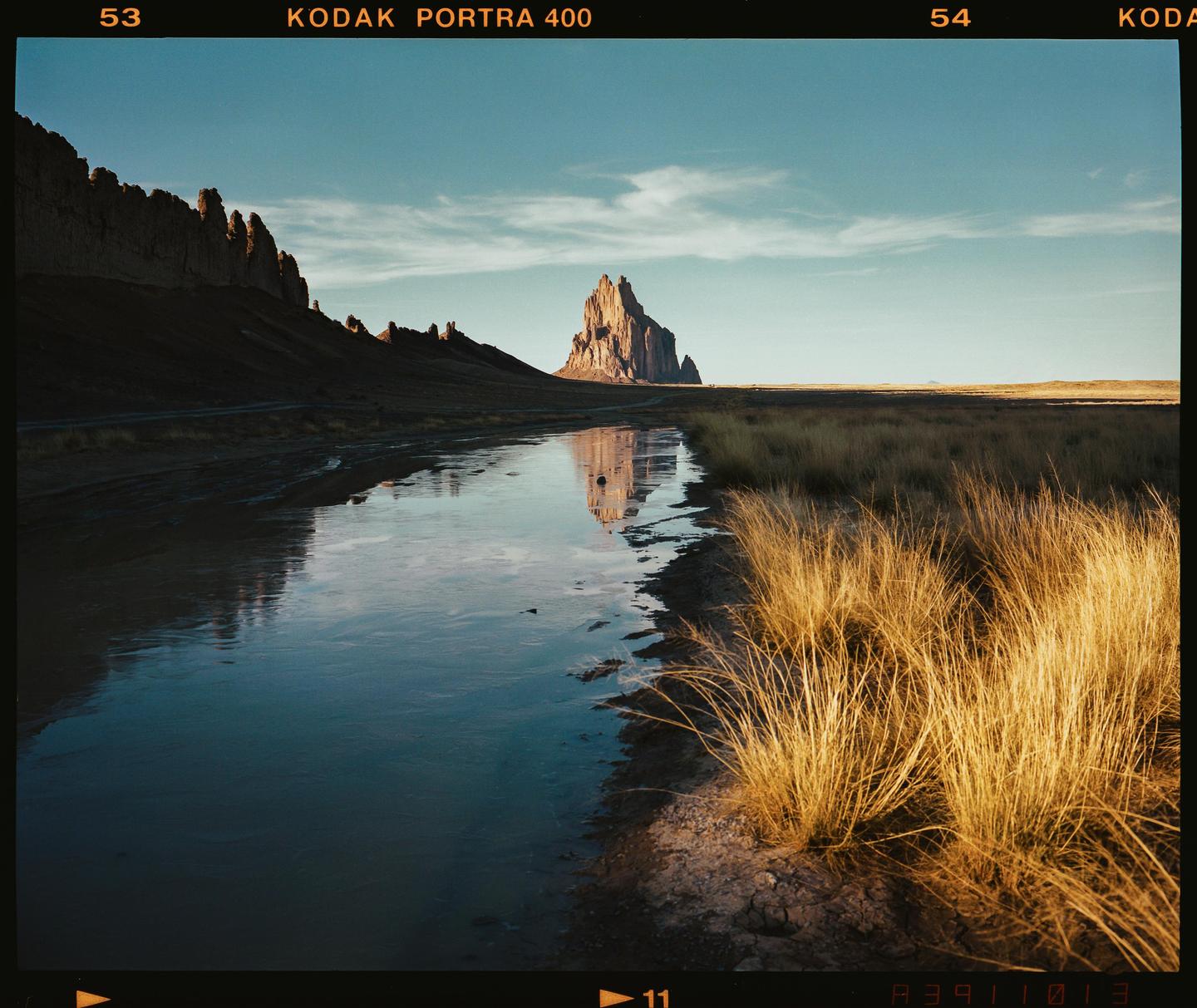 A large rock formation is reflected in a body of water next to a field of dry grass.