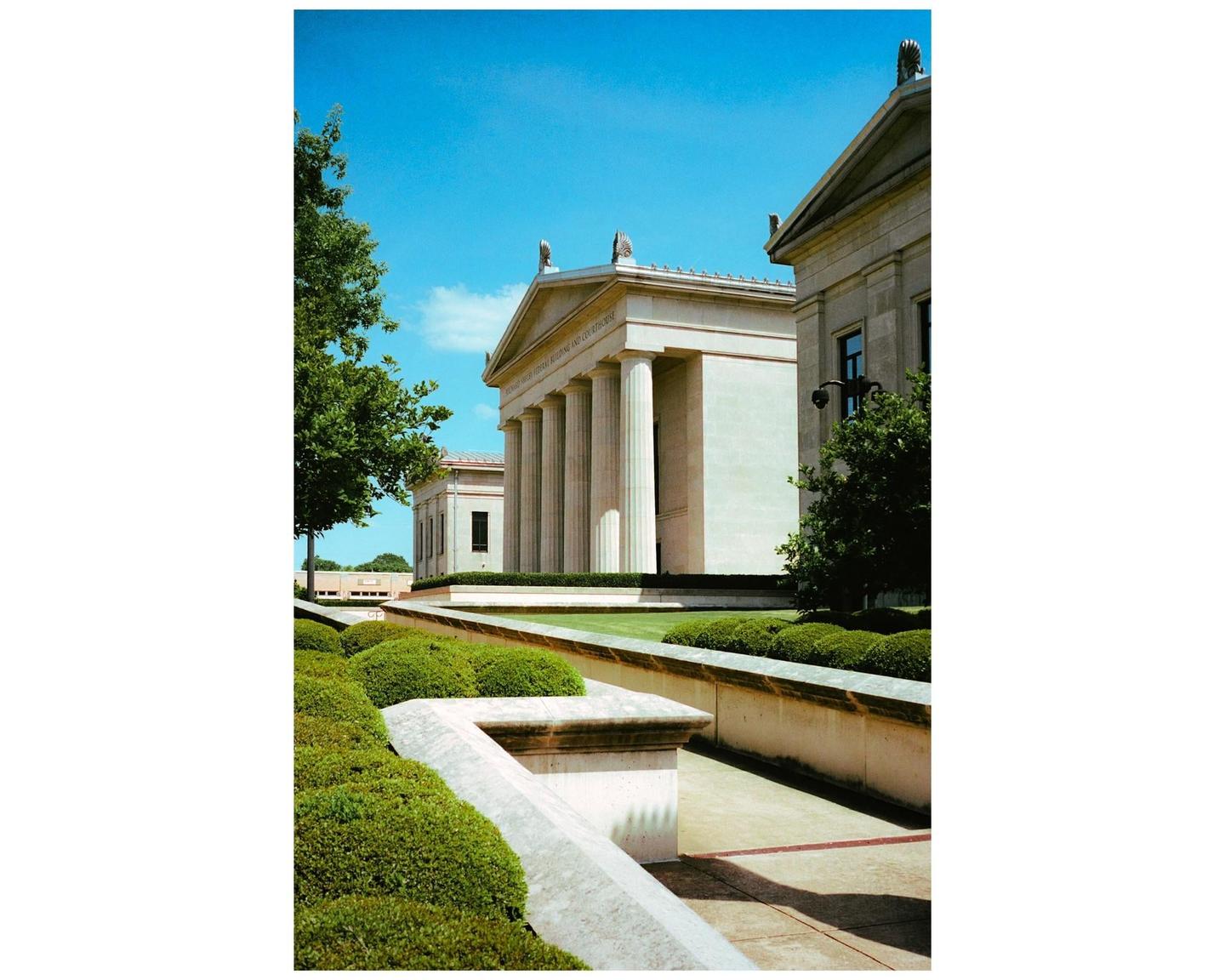 A classical building with columns and a pediment is visible behind a manicured garden and walkway under a clear blue sky.