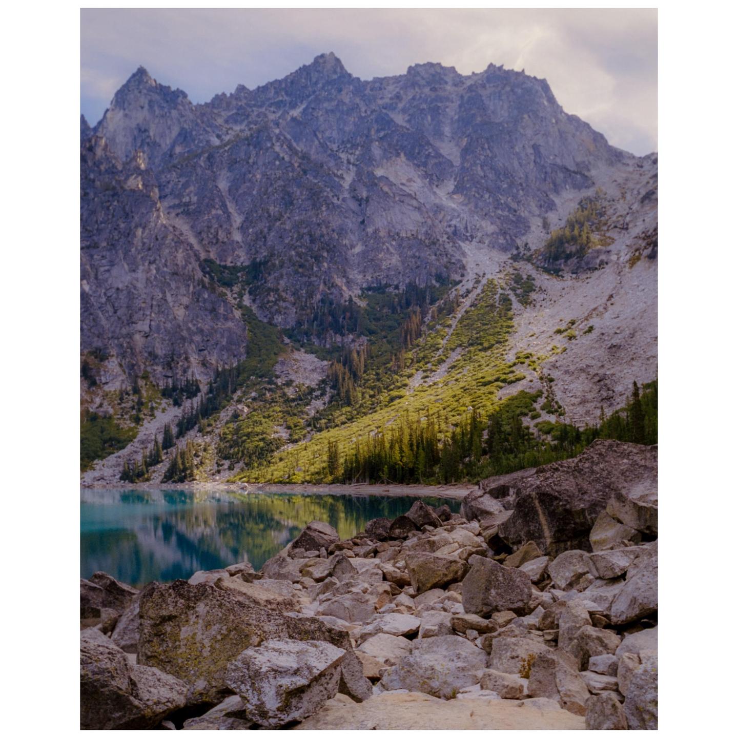 A mountain lake is surrounded by rocky slopes and evergreen trees under a cloudy sky.