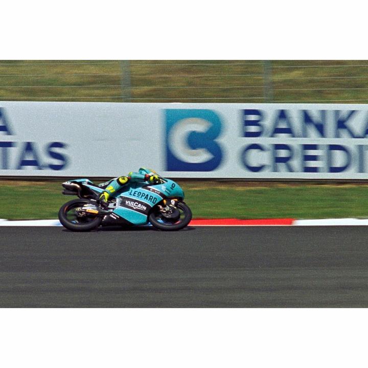 A motorcycle racer leans into a turn on a track with a sponsor banner in the background.