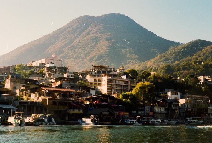 A town with many buildings and boats on the water sits at the base of a large, tree-covered mountain.