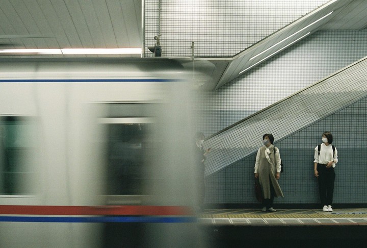 Two women wearing face masks stand on a subway platform as a train passes by.