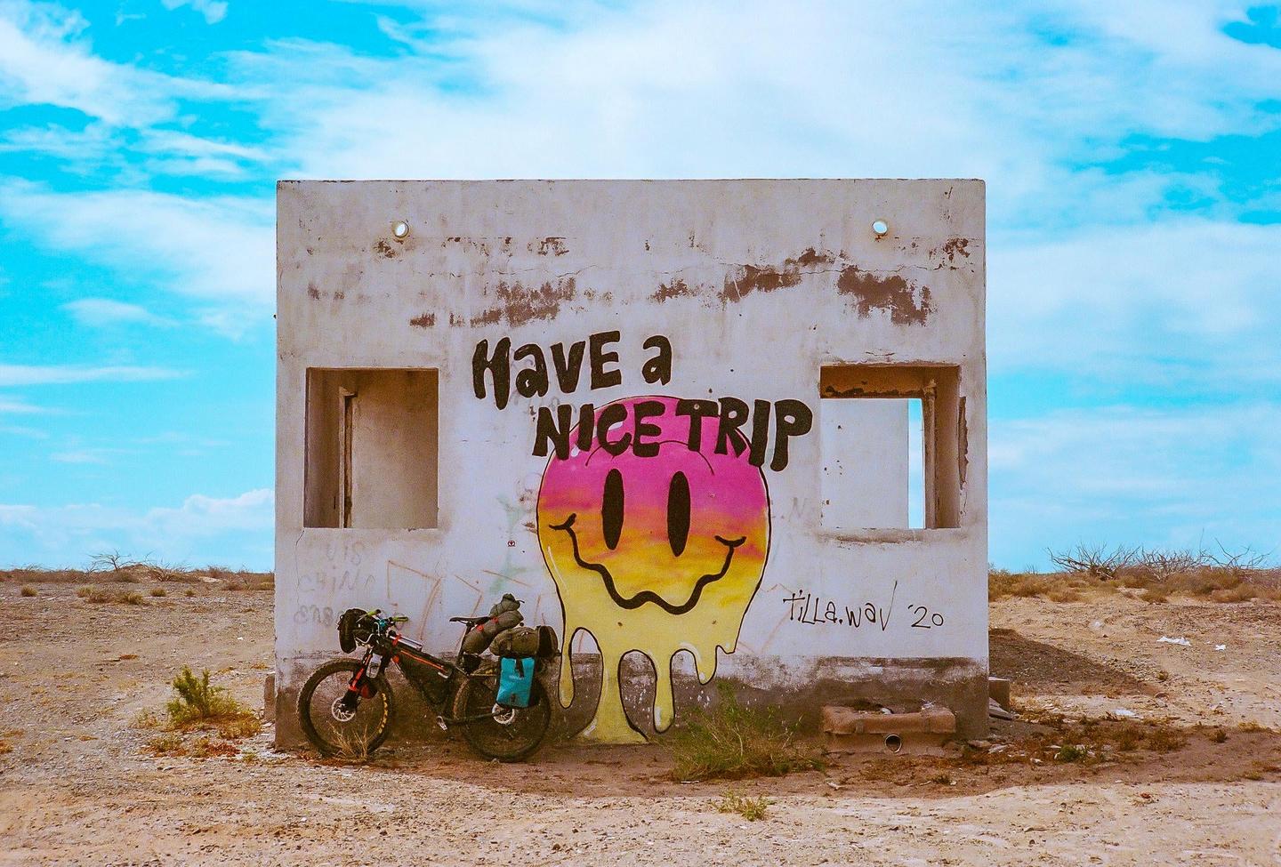 A bicycle is parked in front of a white building with graffiti that says "Have a NICE TRIP" and features a melting smiley face.