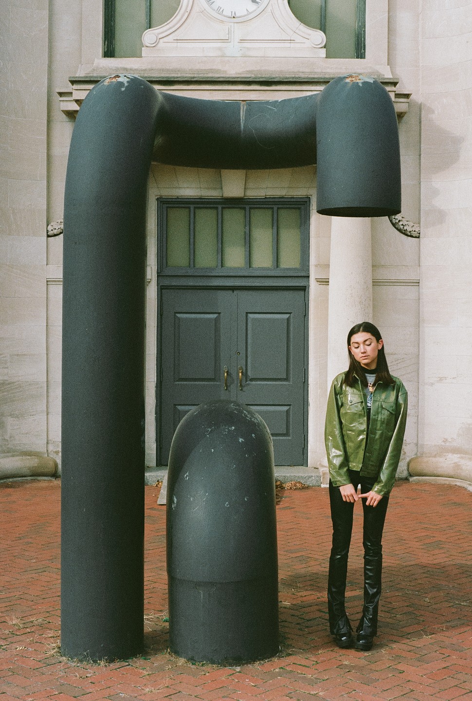 A woman stands next to a large, dark, curved sculpture in front of a building with a clock.
