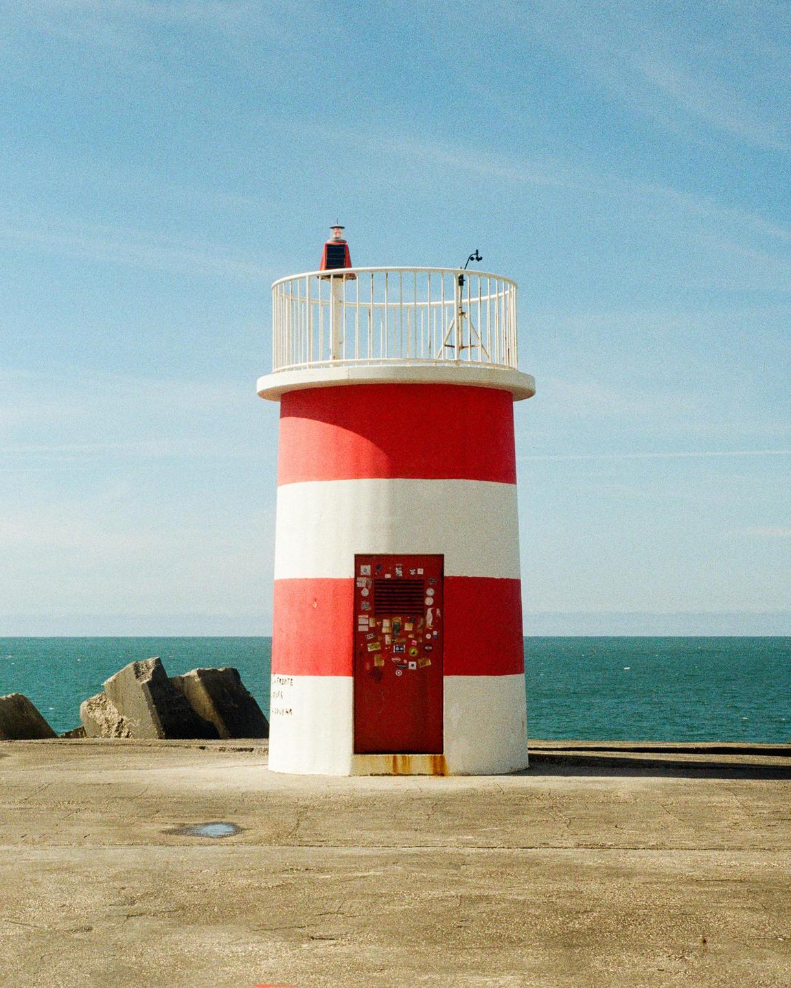 A red and white striped lighthouse stands on a concrete pier next to the ocean under a clear sky.