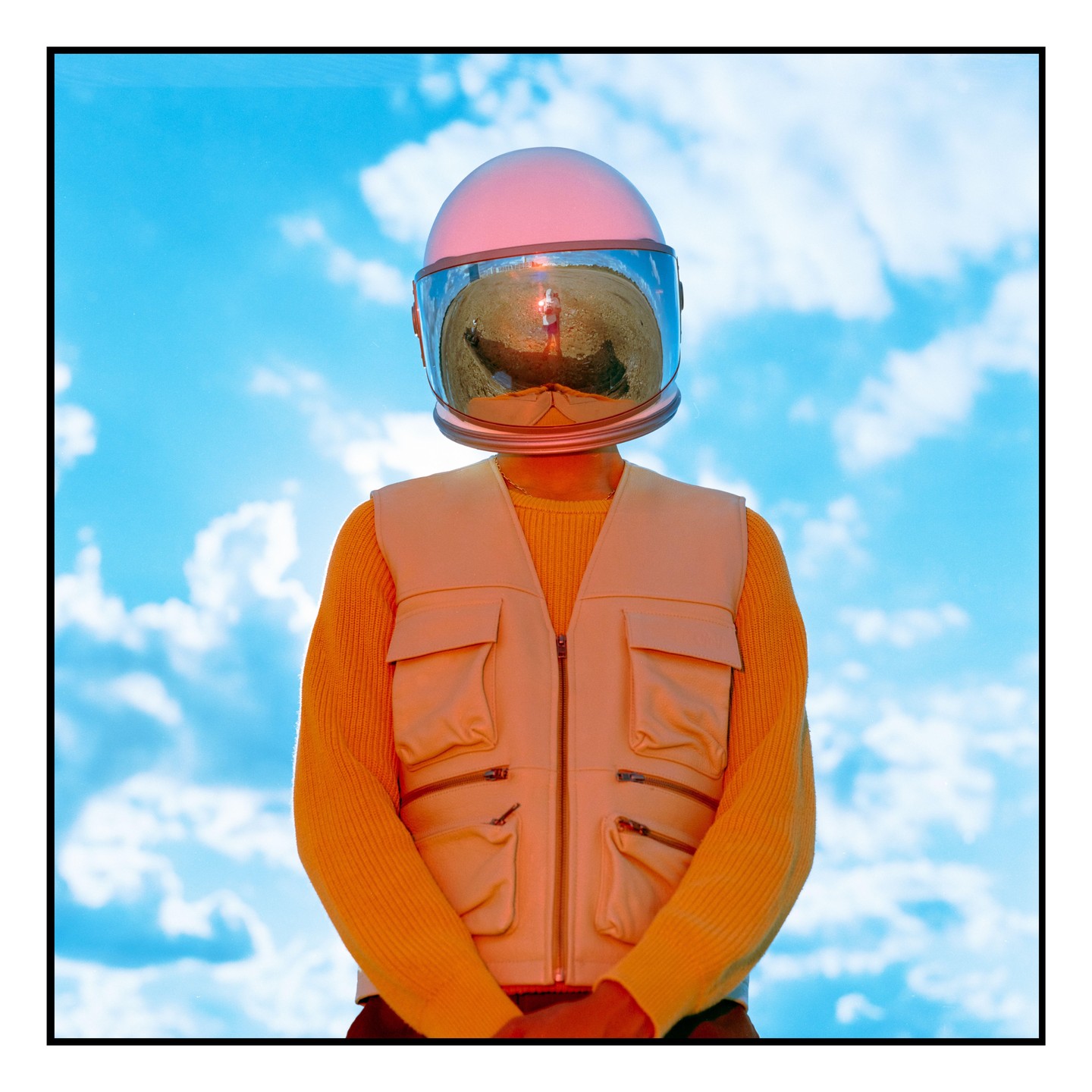 A person wearing a pink astronaut helmet and a cream vest over a yellow sweater stands against a blue sky with clouds.