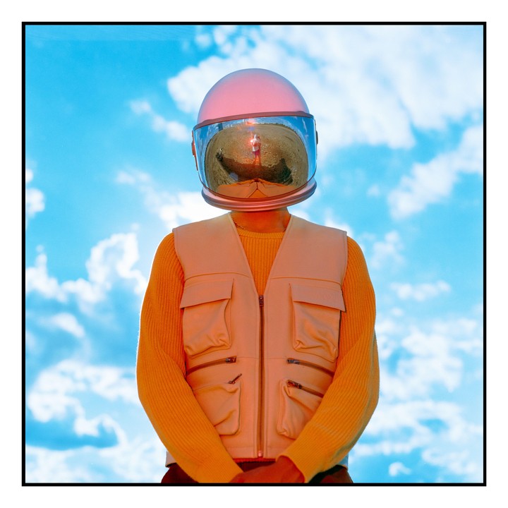 A person wearing a pink astronaut helmet and a cream vest over a yellow sweater stands against a blue sky with clouds.