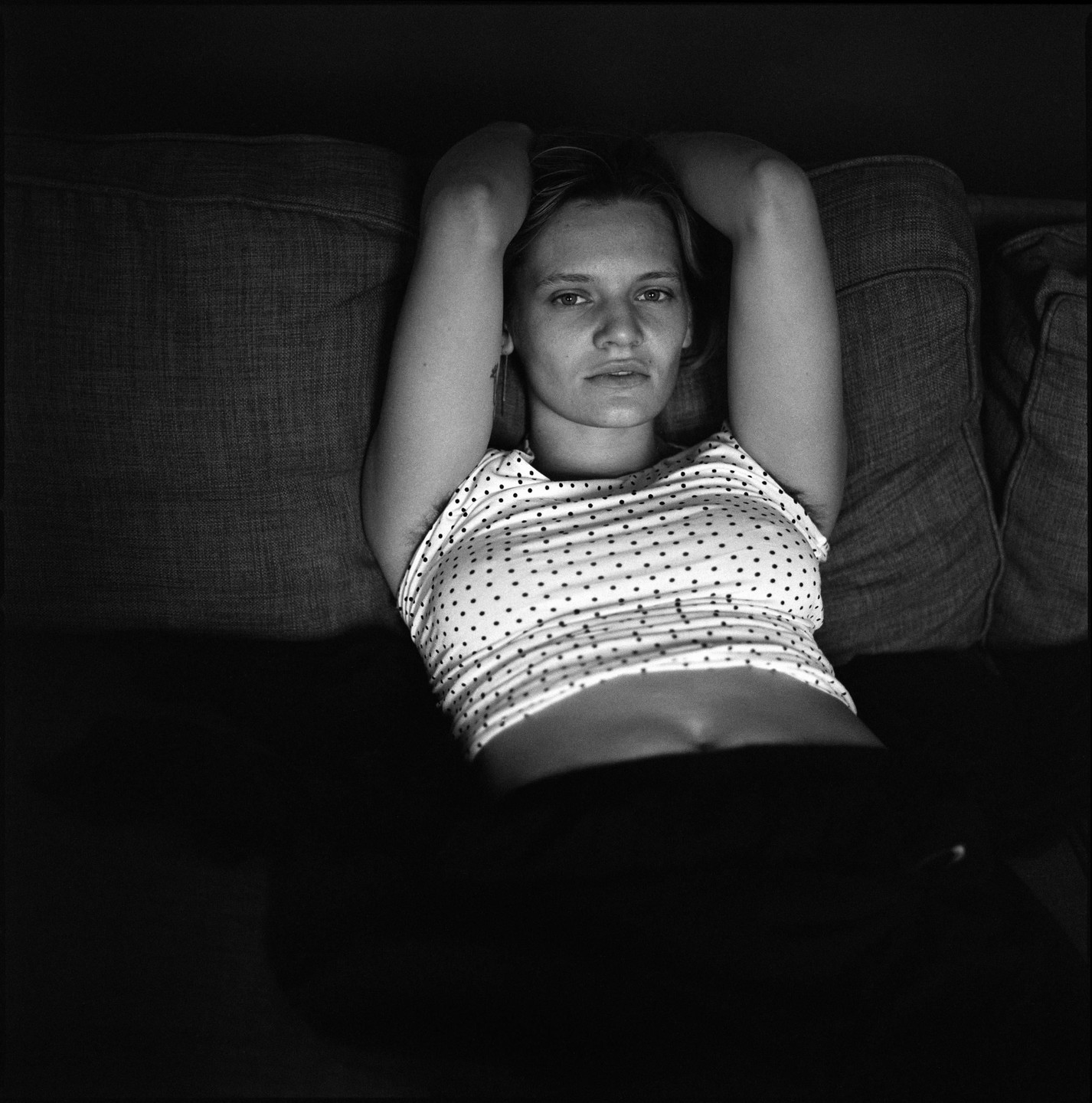 A woman in a polka dot shirt lies on a sofa with her arms behind her head.
