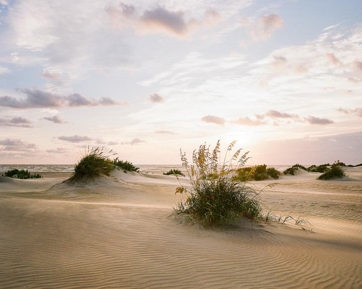 A sandy beach with small dunes and sparse vegetation is illuminated by the setting sun.