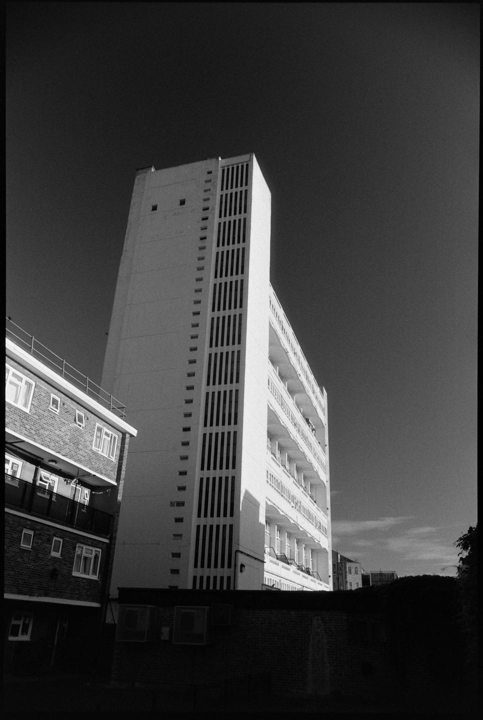 A tall, white building with many windows and balconies stands next to a shorter brick building under a dark sky.