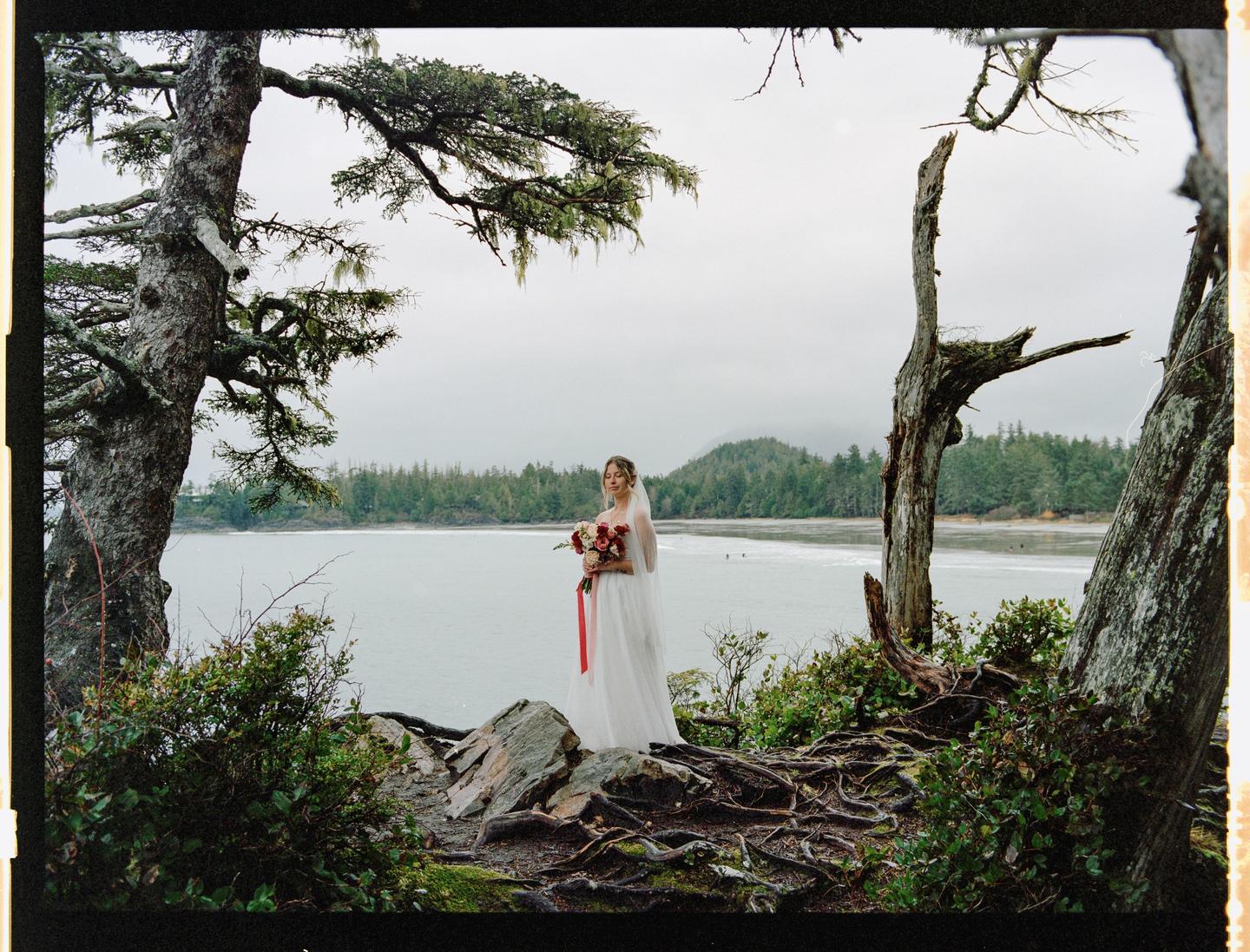A woman in a white dress and veil holds a bouquet of flowers with a red ribbon while standing on a rocky shore with a body of water and forest in the background.