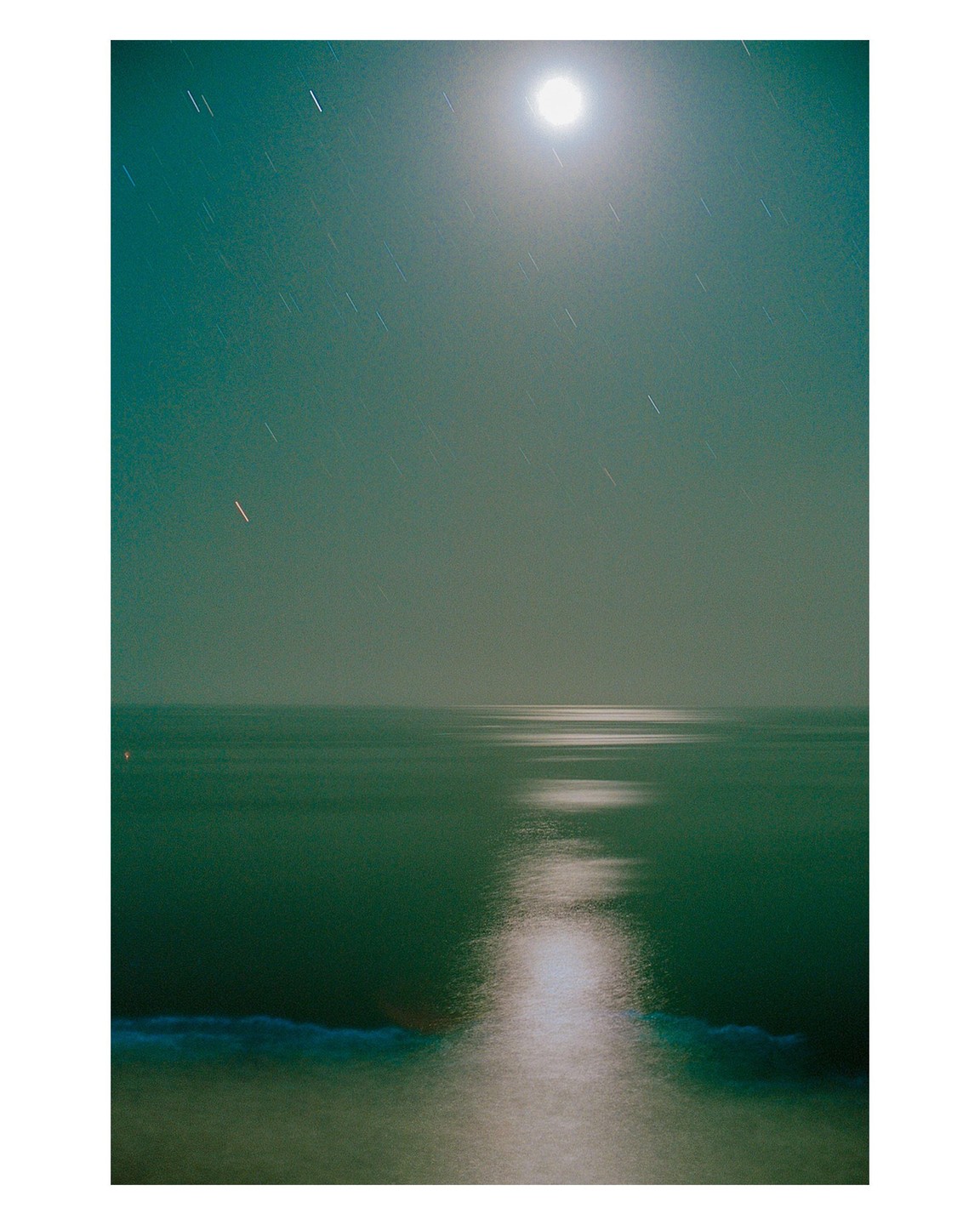 The moon illuminates the ocean and creates a path of light on the water as star trails are visible in the sky.