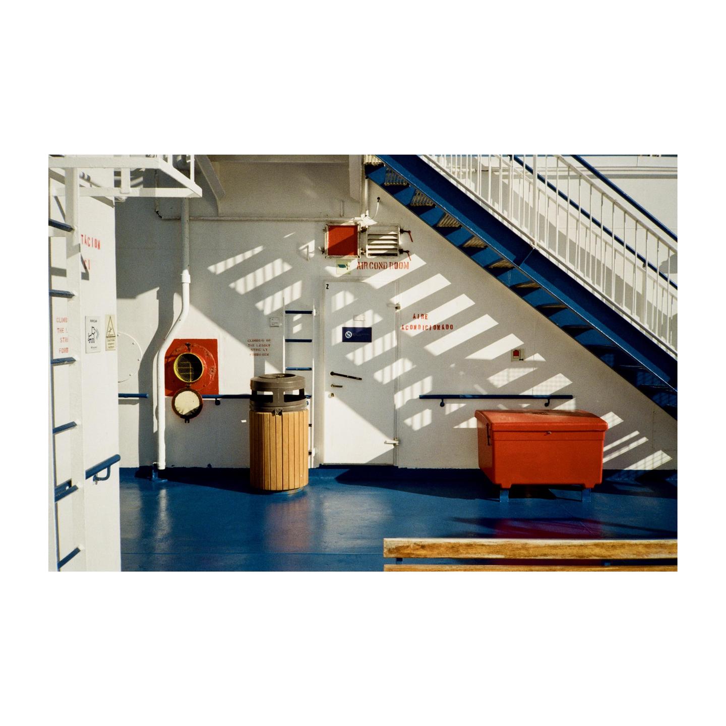 A blue staircase ascends on the right side of a white wall with a red box and a trash can on a blue floor.