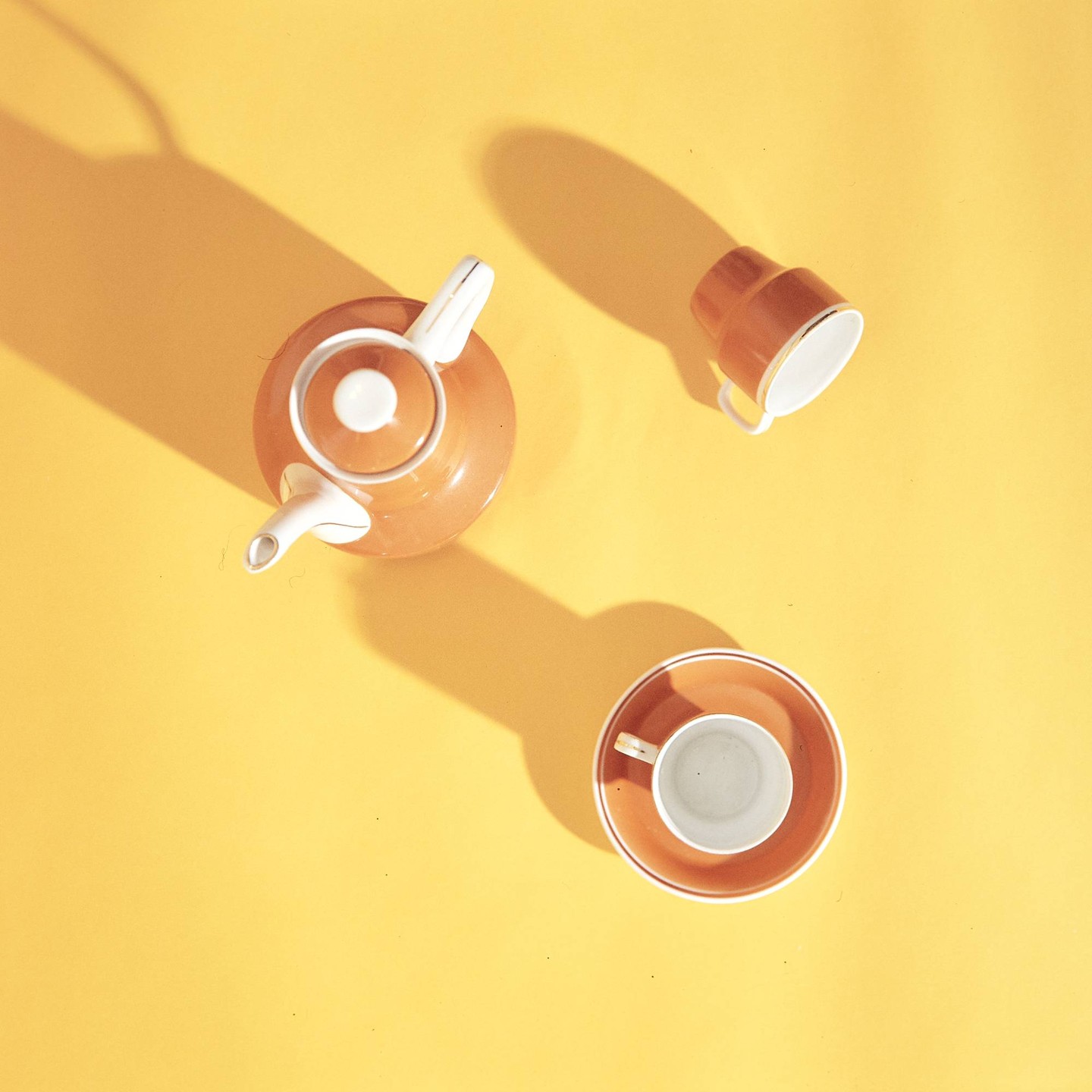A teapot, a cup, and a cup with a saucer are arranged on a yellow surface with shadows.