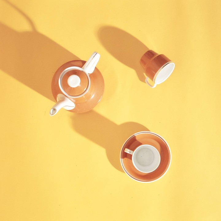 A teapot, a cup, and a cup with a saucer are arranged on a yellow surface with shadows.