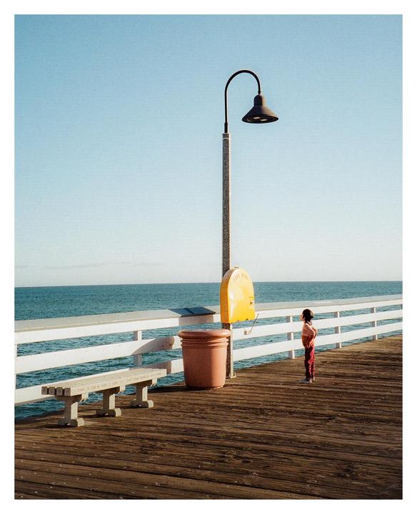 A child stands on a wooden pier looking out at the ocean under a clear sky.