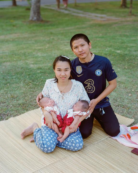 A couple sits on a mat in a grassy park with two sleeping babies.
