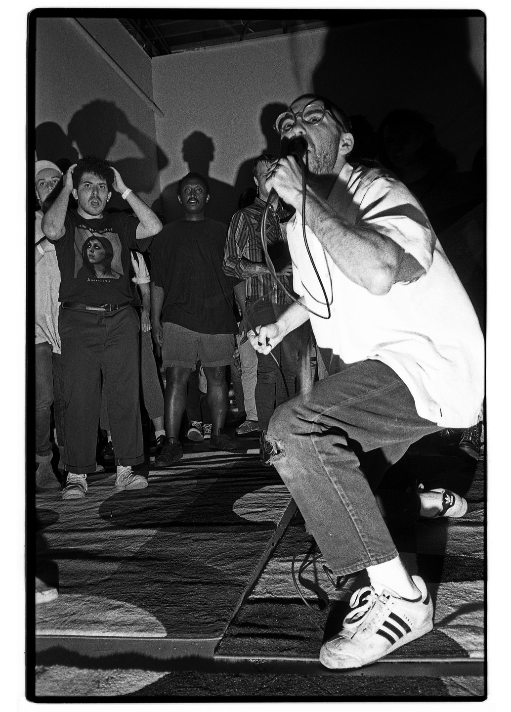 A man wearing glasses and ripped jeans sings into a microphone on a stage with other men standing behind him.