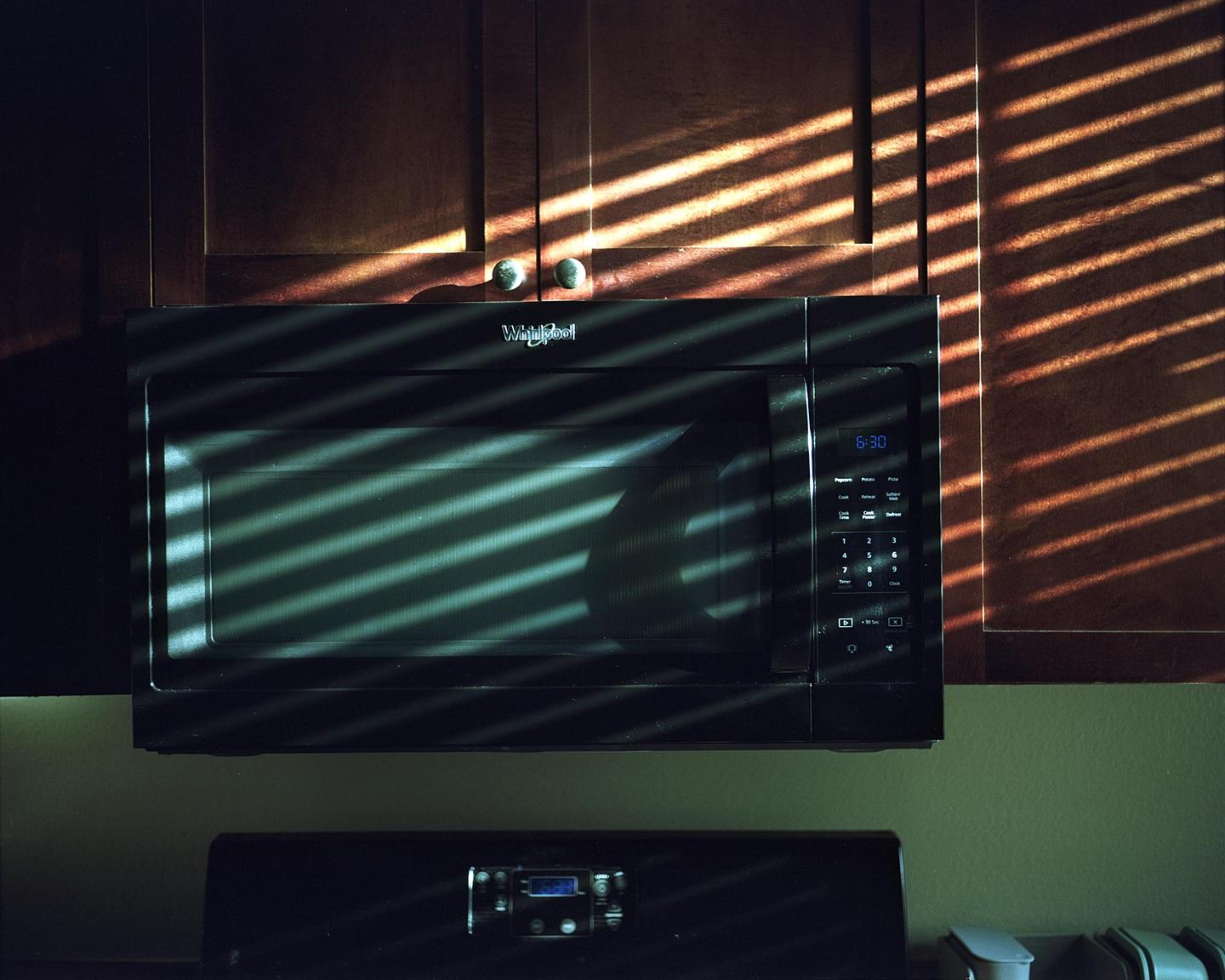 Sunlight streams through blinds, casting striped shadows across a microwave and kitchen cabinets.