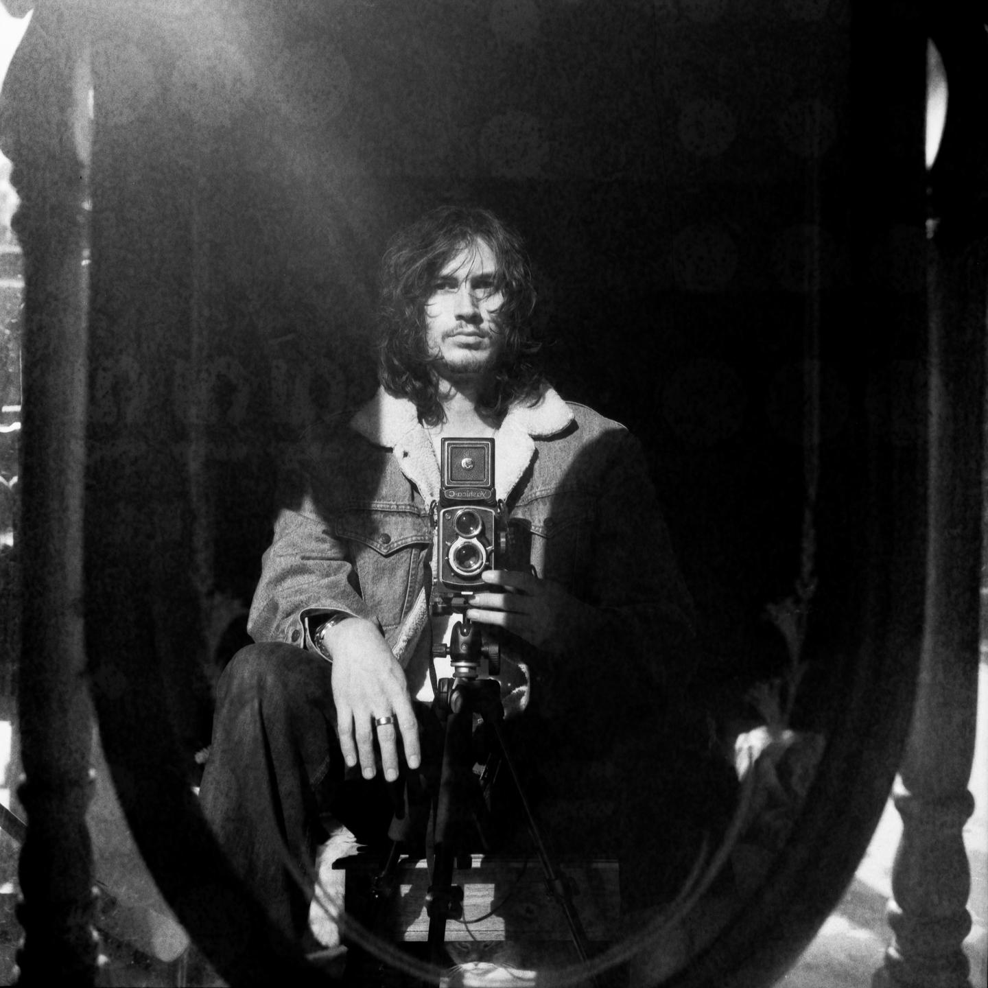 A man with long hair and a denim jacket holds a camera on a tripod in a mirror self-portrait.
