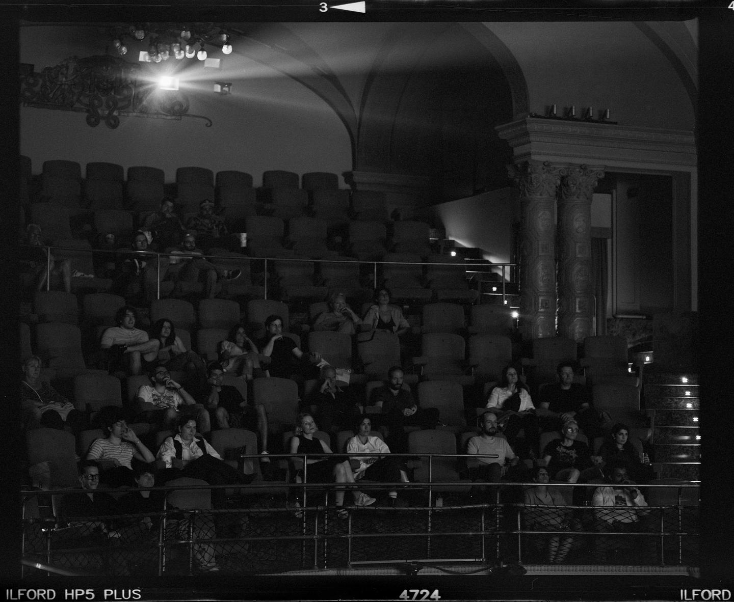 A black and white photo shows an audience watching a film in a dark theater.