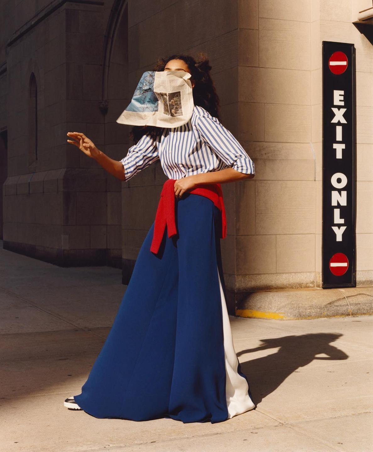 A woman with a newspaper covering her face stands on a sidewalk next to a building and an "exit only" sign.