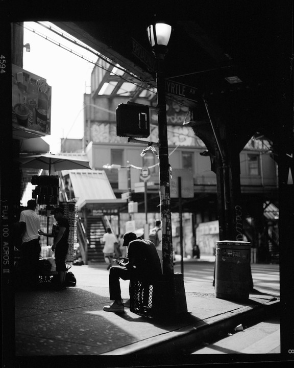A man sits on a crate on a sunlit street corner under a street sign for Myrtle Avenue and Broadway.