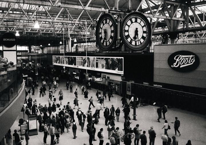 A large crowd of people walk through a train station with two large clocks hanging above them.