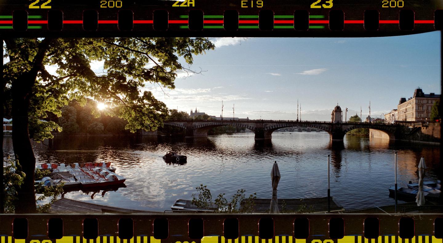 The sun sets over a river with a bridge and buildings in the distance, framed by trees and film sprockets.