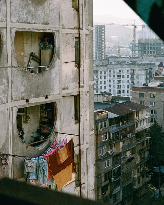 Laundry hangs on a clothesline outside a concrete building with circular windows overlooking a city.