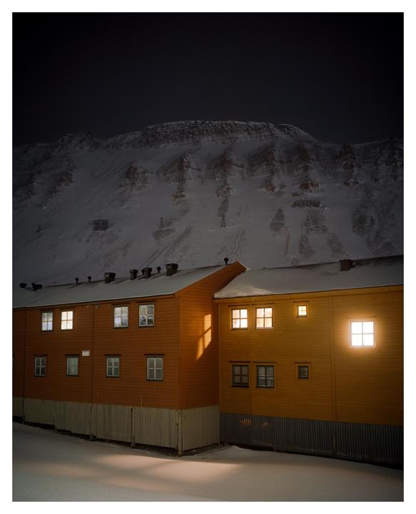 A yellow building with illuminated windows stands in the snow at night, with a large snow-covered mountain in the background.