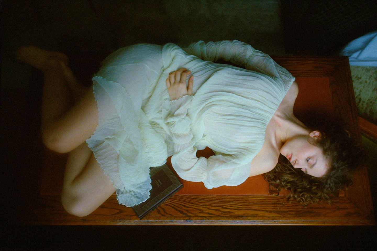 A woman in a white dress lies on a wooden surface with a book next to her.