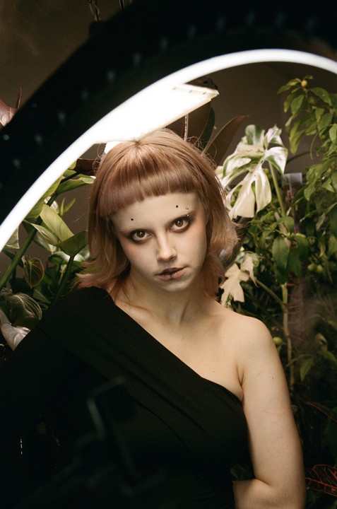 A woman with short bangs and dark makeup looks directly at the viewer, surrounded by plants and a bright ring light.