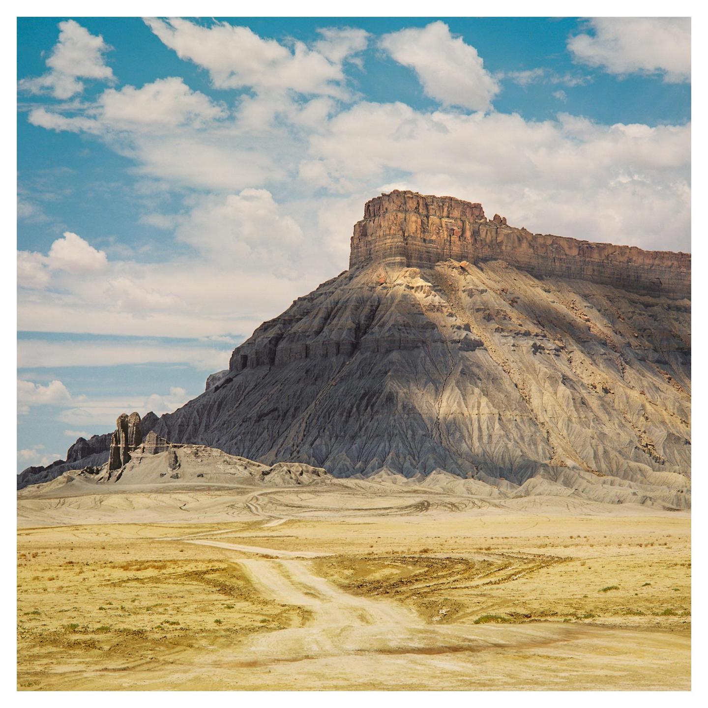 A dirt road leads to a large mesa under a blue sky with white clouds.