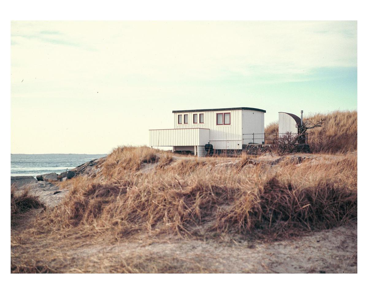 A white house with red window frames sits on a grassy dune overlooking the ocean.