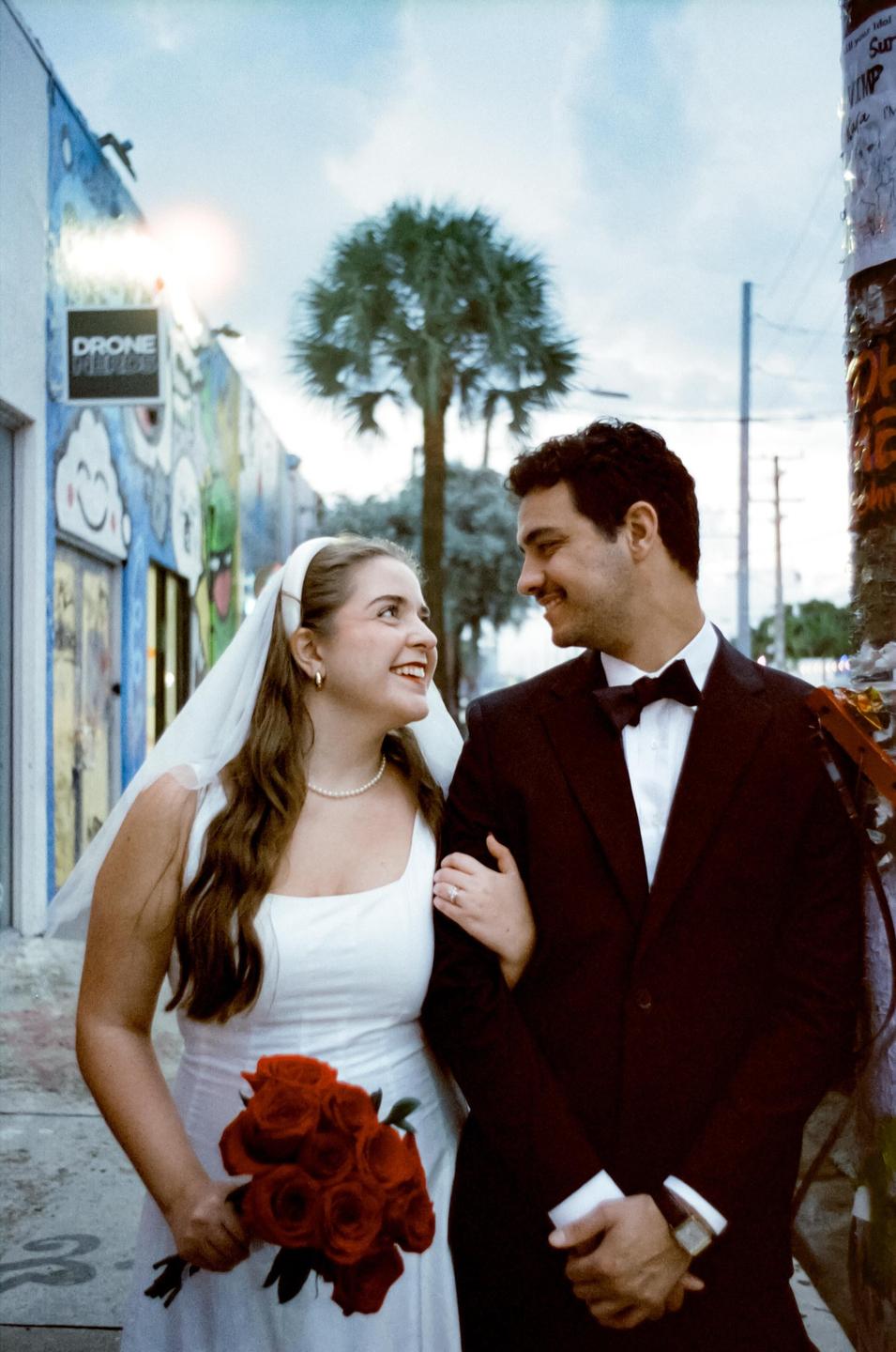 A couple in wedding attire stands on a street with a colorful mural and a palm tree in the background.
