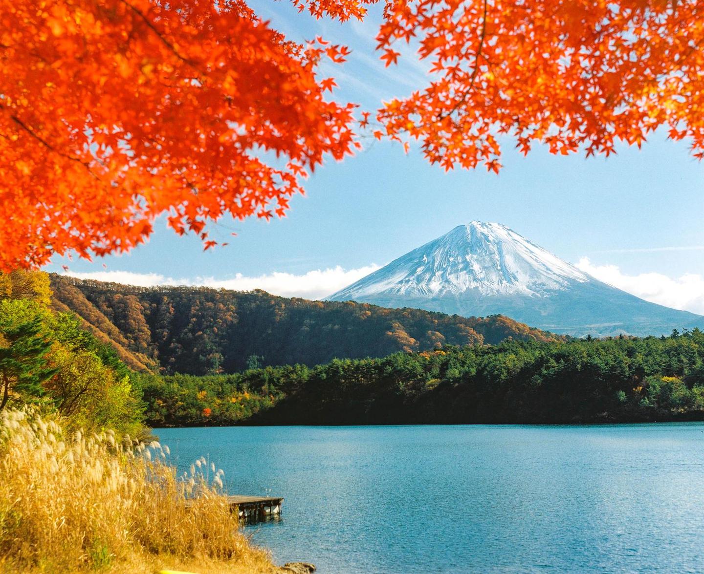 A snow-capped mountain rises behind a lake surrounded by autumn foliage and a small wooden dock.