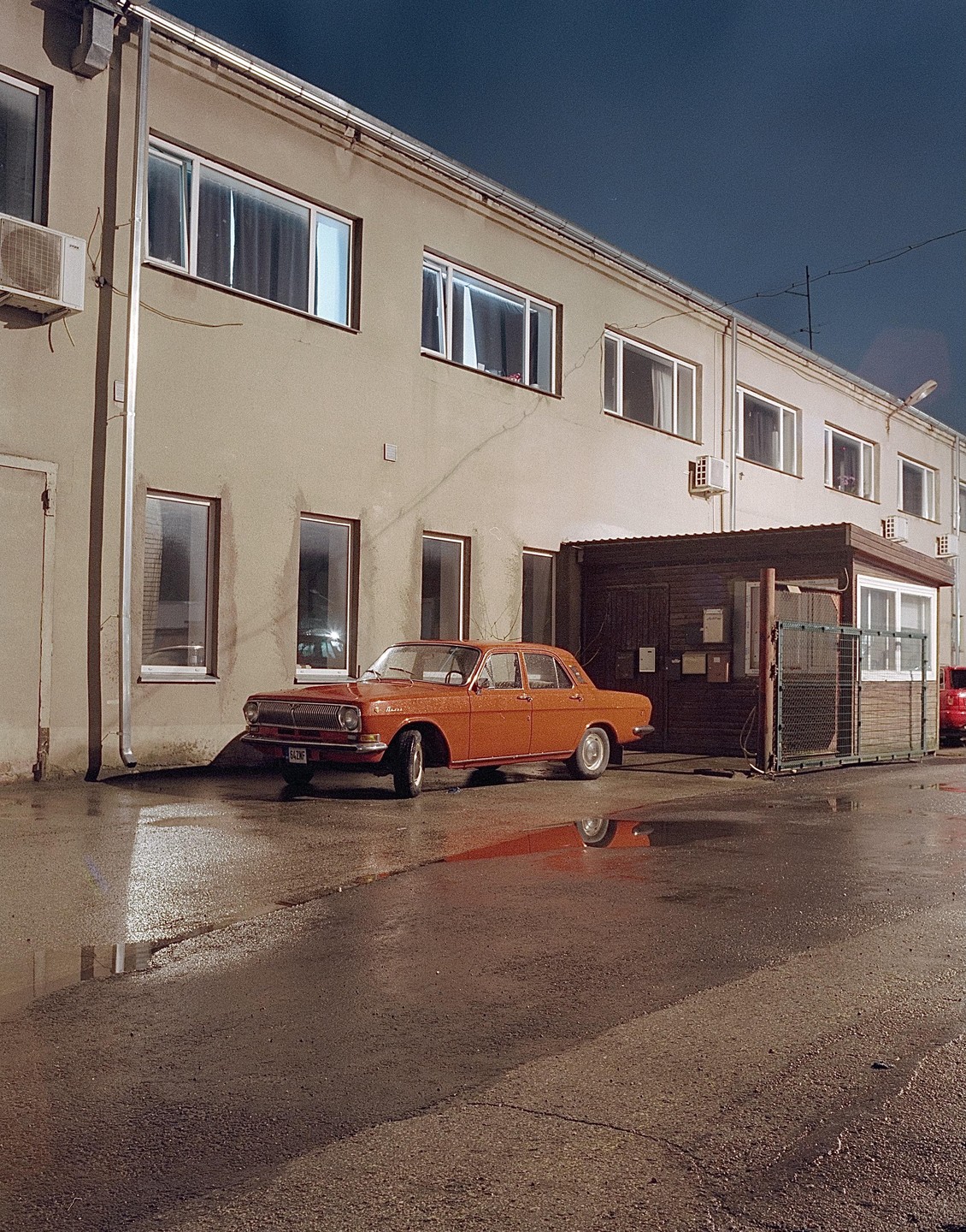 An orange car is parked on a wet street next to a building at night.