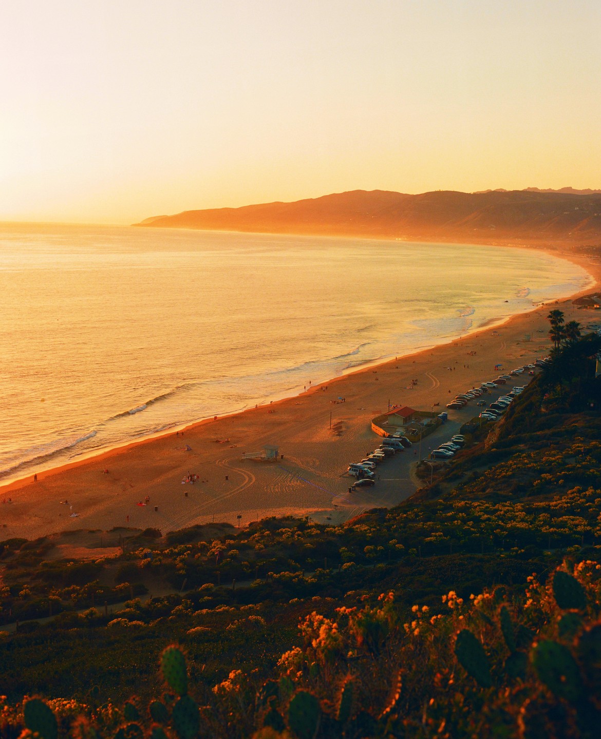 An aerial view shows a beach with a parking lot and mountains in the distance during sunset.