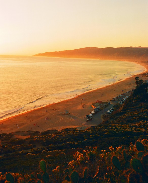 An aerial view shows a beach with a parking lot and mountains in the distance during sunset.