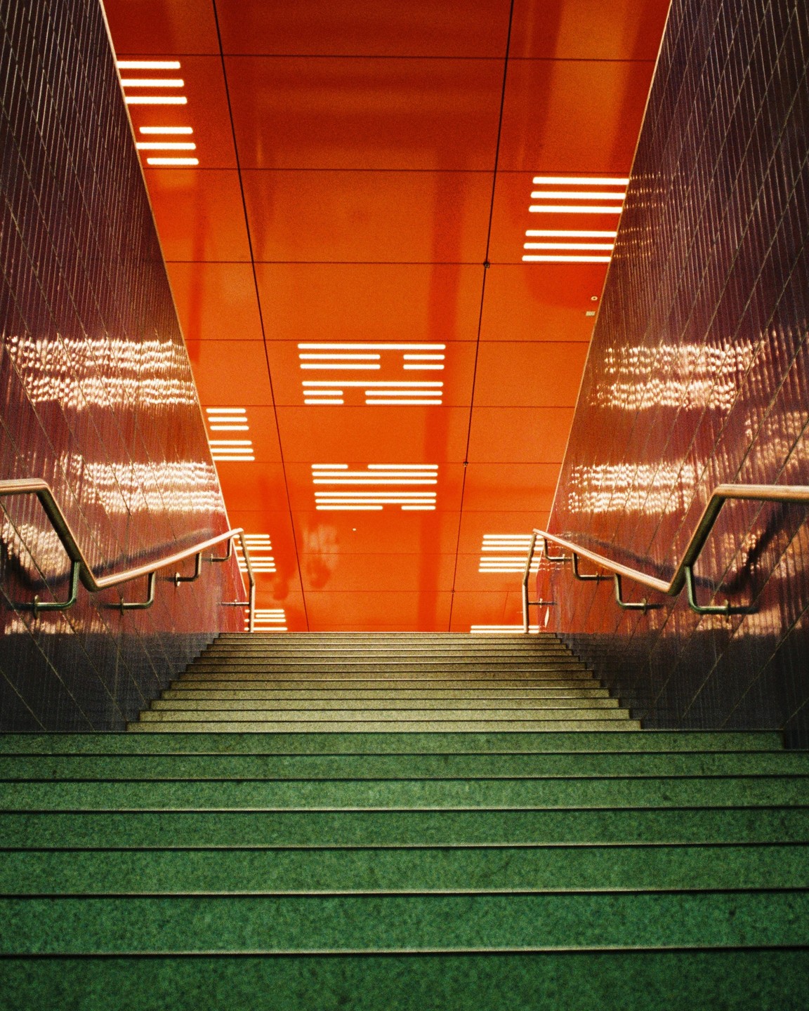 A long staircase with green steps leads up to a bright orange ceiling with rectangular lights.