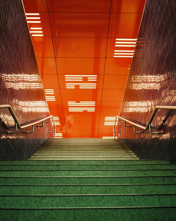 A long staircase with green steps leads up to a bright orange ceiling with rectangular lights.