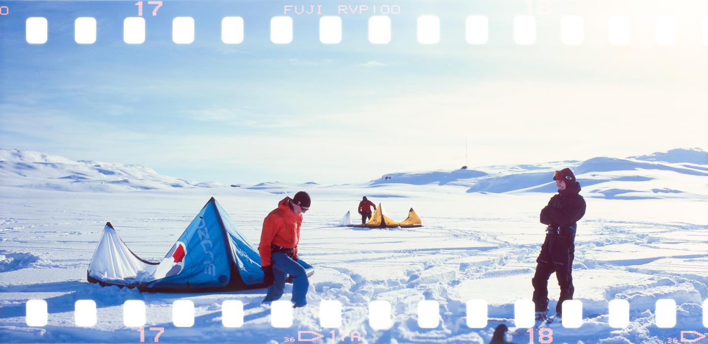 Three men are on a snow-covered field with kites, surrounded by snowy hills under a bright sky.