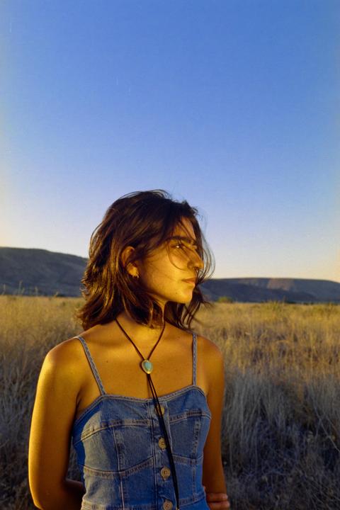 A woman wearing a denim dress and bolo tie stands in a field of dry grass with mountains in the background.