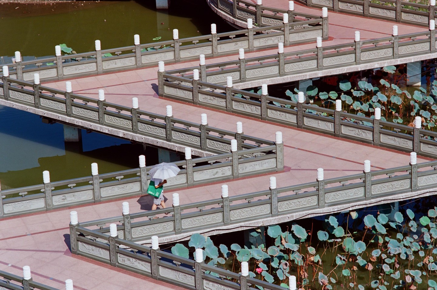 A person with an umbrella walks along a zig-zag bridge over a pond with lily pads.