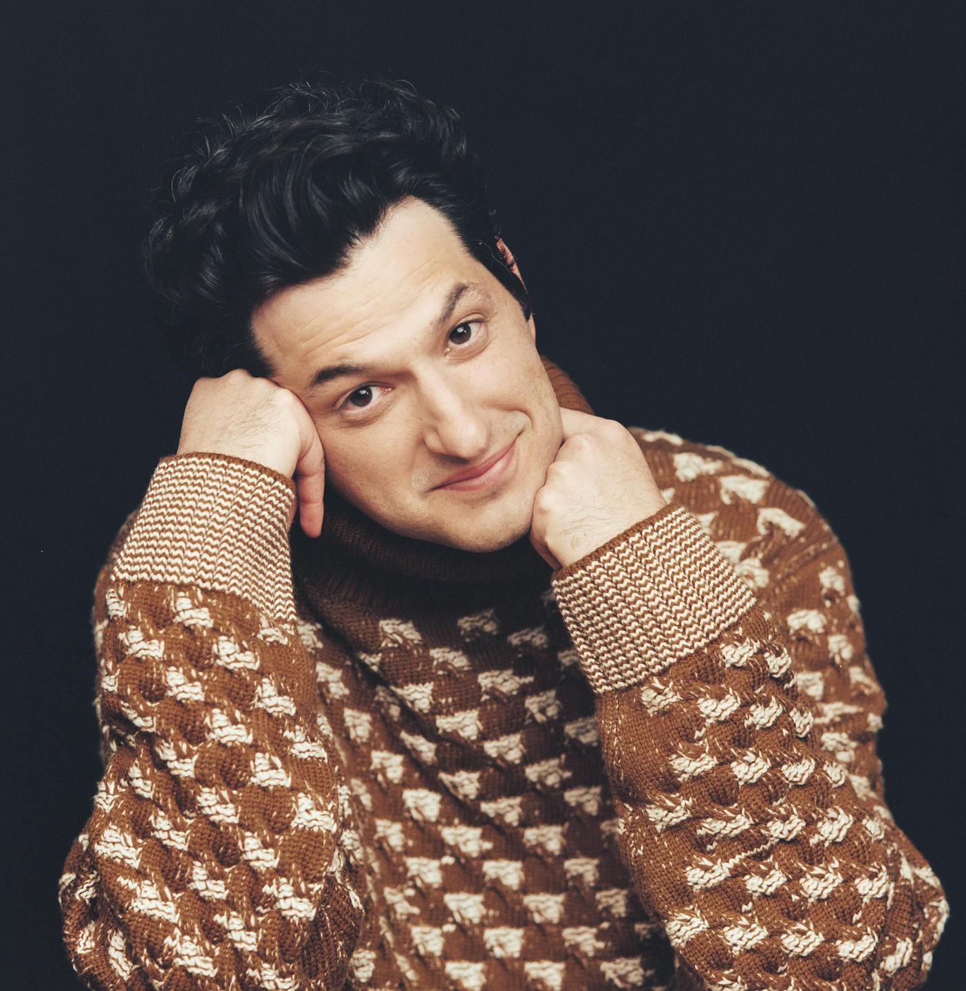 A man in a brown patterned sweater rests his chin on his hands and smiles at the viewer.