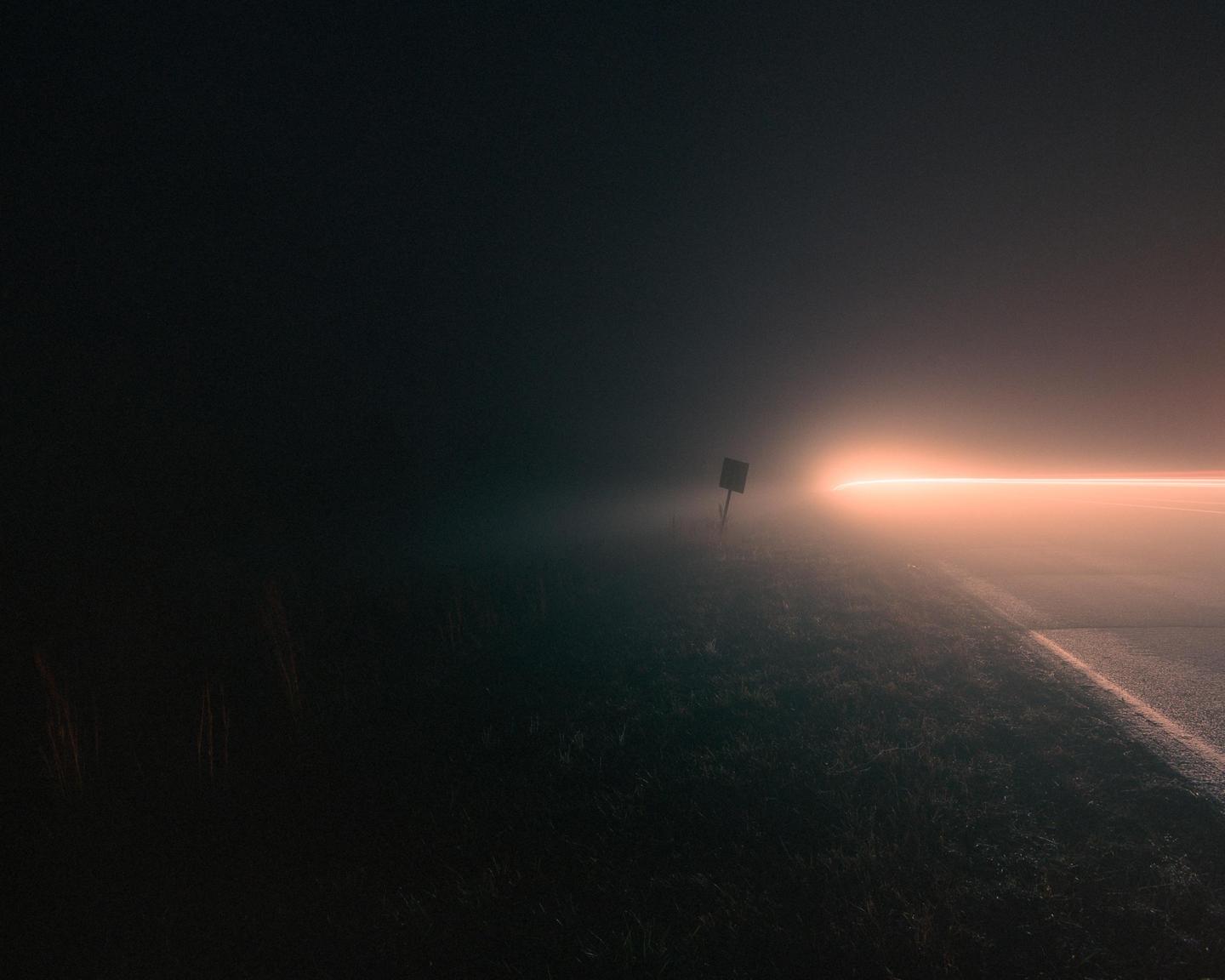A light trail from a vehicle illuminates a foggy road at night.