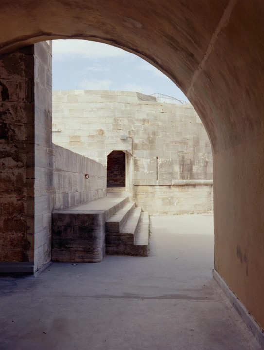 A stone archway frames a view of a stone wall with a small doorway and a set of steps.